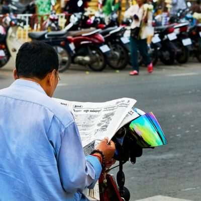 A man on a stationary motorbike reading a newspaper on the street in Cambodia.