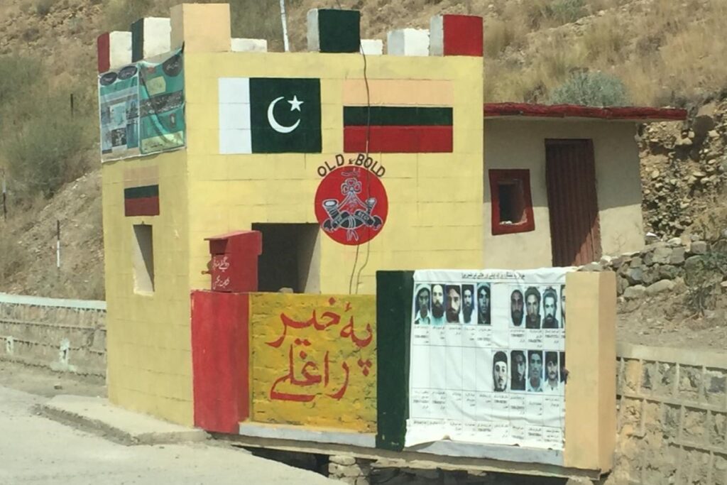Military checkpoint along a road in North Waziristan. A red sign reads Welcome in Pashto, and wanted militant posters are displayed beside it (August/2019).